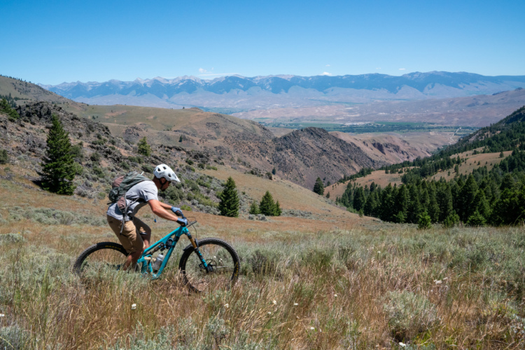 A mountain biker navigates through a grassy hillside with rocky terrain, wearing a helmet and a backpack. In the background, expansive mountains and a clear blue sky create a picturesque landscape. The scene captures the thrill of outdoor biking in a scenic environment.