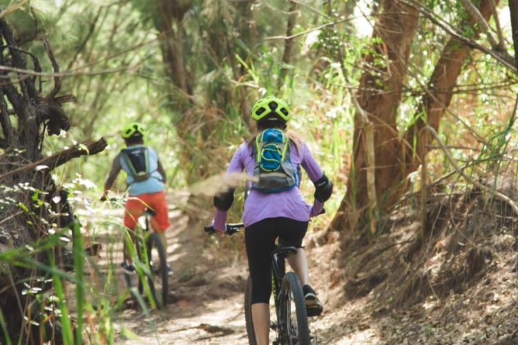 Two cyclists ride on a narrow trail through a wooded area, surrounded by greenery. One cyclist wears a purple shirt and black pants, while the other has a gray shirt and orange shorts. Both are wearing bright green helmets and carrying backpacks, as they navigate the outdoor path.