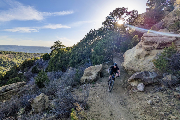 A mountain biker riding along a narrow dirt trail surrounded by rocky terrain and sparse vegetation, with the sun setting in the background and illuminating the landscape.
