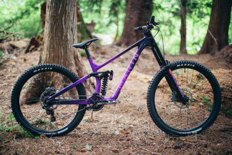 A purple mountain bike with a sleek design is positioned on a forest floor covered in pine needles, surrounded by trees. The bike features prominent suspension, wide tires, and a modern frame, highlighting its suitability for trails and rugged terrain.