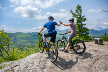 Two mountain bikers share a moment of camaraderie on a rocky ledge overlooking a lush, green valley and distant mountains. They are wearing helmets and casual biking attire, with one reaching out to fist bump the other. The sky is partly cloudy, and the scene conveys a sense of adventure and friendship in the outdoors. Blueberry Hill mountain bike trail.
