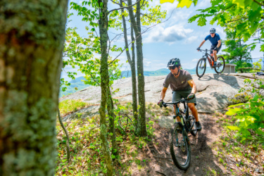 Two mountain bikers navigate a rocky trail surrounded by trees and greenery. The scene features bright blue skies and scenic mountains in the background, showcasing an active outdoor adventure in a natural setting. Blueberry Hill mountain bike trail.