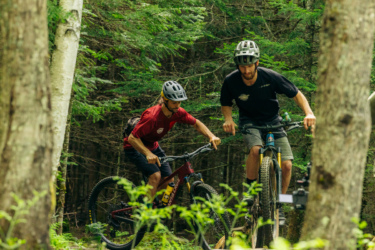 Two mountain bikers navigating through a forested trail, focused on their ride. One rider is in a red shirt, while the other is wearing a black shirt, both wearing helmets. The scene captures the lush greenery of the forest and the challenging terrain they are tackling. Lussi & Loggers mountain bike trail.