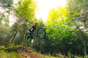 A mountain biker mid-jump over a rocky terrain in a dense forest with lush green trees and bright foliage above. Lussi & Loggers mountain bike trail.