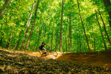 A mountain biker leans into a turn on a dirt trail surrounded by lush green trees, with sunlight filtering through the leaves. The trail is covered in fallen leaves, capturing the energetic action of the sport in a vibrant forest setting. East Branch Community Trails mountain bike trail.