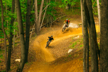 Two mountain bikers navigate a winding dirt trail in a lush green forest. One rider leans into a turn, while the other is airborne, showcasing their skills amidst tall trees and scattered leaves on the ground. East Branch Community Trails mountain bike trail.