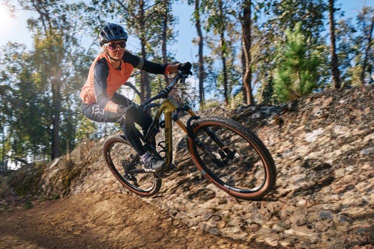 A mountain biker in an orange and black outfit performs a jump on a rocky trail surrounded by trees in a sunlit forest. The rider wears a helmet and sunglasses for protection, showcasing an action-packed moment in outdoor cycling.