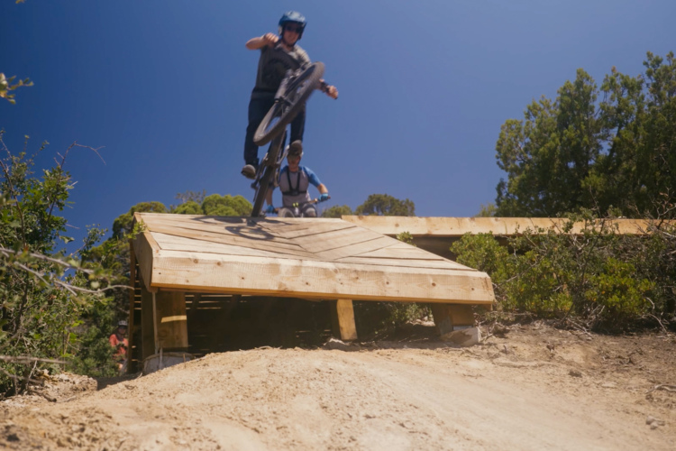 A mountain bike rider mid-jump off a wooden ramp on a sunny day, with another rider in the background. The setting features a dirt trail surrounded by greenery under a clear blue sky.