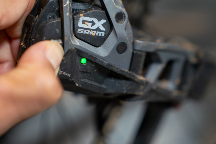 Close-up of a hand pressing a SRAM GX bike shifting lever, showing a green indicator light, with dirt visible on the lever, suggesting outdoor use.