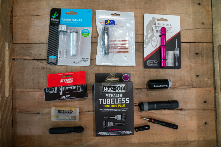 An assortment of bike tire repair kits and tools displayed on a wooden surface. Items include a tubeless tackle kit, a puncture repair kit, and various puncture plugs from brands like Muc-Off and Stan's. The collection showcases tools for removing valve cores and repairing punctures, designed for maintaining tubeless tire setups.