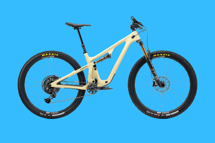 A modern mountain bike with a light beige frame, featuring a full suspension design, wide tires with a Maxxis tread, and a prominent front fork. The bike is showcased against a bright blue background.