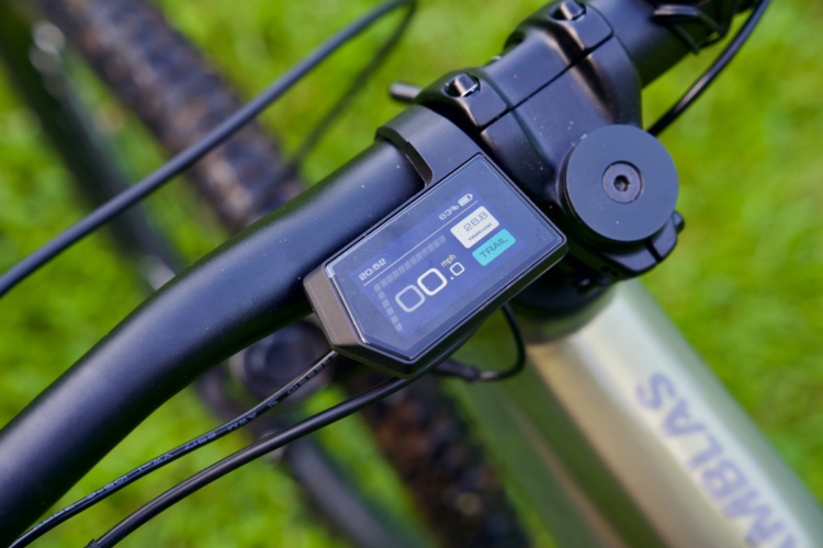 Close-up view of a bicycle handlebar with a digital display showing speed and 63% battery life, set against a background of green grass. The display reads 00.0 mph, with additional information visible, indicating features related to bike performance.