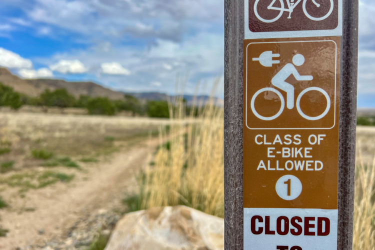 Sign indicating e-bike class allowed on a biking path, with a blurred natural landscape and cloudy sky in the background. The sign specifies allowed classes and indicates the area is closed to certain users.