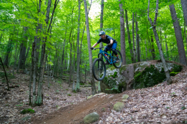 A mountain biker in protective gear jumps off a rock while riding through a forest trail, surrounded by lush green trees and a carpet of leaves on the ground. Hardy Road mountain bike trail.