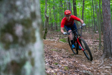 A mountain biker in a red jersey and helmet maneuvers a winding trail through a lush green forest, surrounded by trees and rocky terrain. Hardy Road mountain bike trail.
