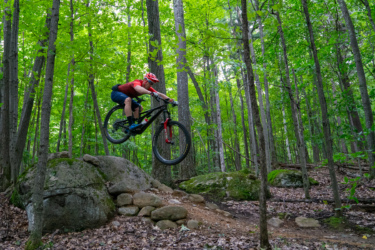 A mountain biker in a red shirt and helmet jumps off a rock ledge in a lush green forest, surrounded by tall trees and foliage. The bike is airborne, showcasing dynamic movement and excitement in an outdoor adventure setting. Hardy Road mountain bike trail.