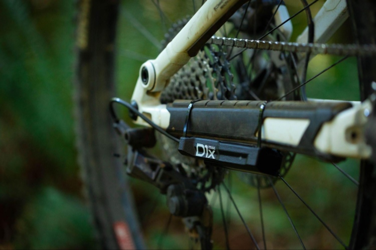 Close-up of a mountain bike's drivetrain, showcasing the gear cassette, chain, and portion of the frame. The image highlights a black D1x Trail component attached to the bike, set against a blurred green background of foliage.