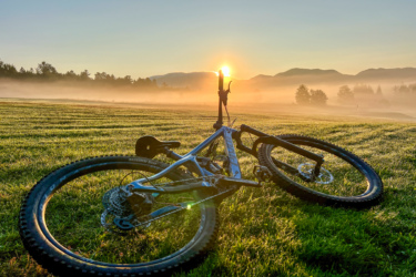 A mountain bike resting on vibrant green grass, with the sun rising in the background, casting a warm glow over misty mountains. The scene captures the peacefulness of an early morning outdoors, surrounded by nature. Lussi & Loggers mountain bike trail.