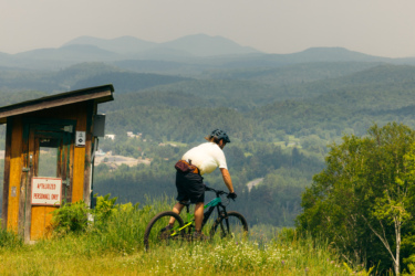A mountain biker pauses on a scenic overlook, facing a vast mountainous landscape with layers of green hills and distant peaks. In the foreground, a small wooden structure displays a sign reading "Authorized Personnel Only." The biker, wearing a helmet and riding gear, stands beside their bike amidst tall grass and wildflowers, enjoying the view. Mount Pisgah Ski Area mountain bike trail.