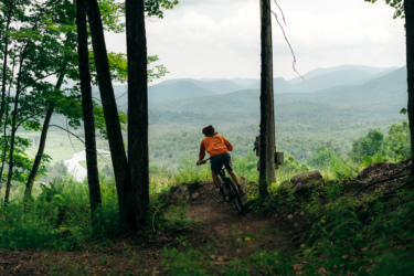 A person riding a mountain bike along a dirt trail, surrounded by trees and overlooking a scenic landscape with mountains and a river in the background, on a cloudy day. Mount Pisgah Ski Area mountain bike trail.