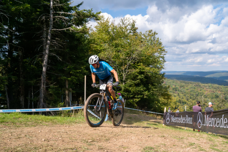 A mountain biker in a blue jersey and helmet navigates a dirt trail surrounded by trees, leaning into a turn with one foot raised. The background features a scenic view of hills and a cloudy sky. Spectators can be seen near a banner on the right side.