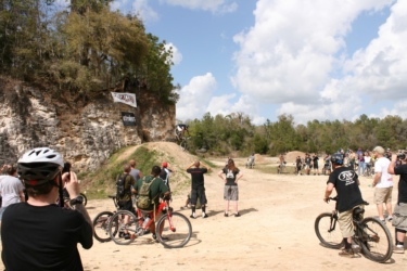 A group of spectators watches a mountain biker performing a jump off a ramp at a dirt biking event. The scene is set outdoors with a rocky cliff in the background, surrounded by trees and blue sky with fluffy clouds. Several spectators are taking photos and cheering, while others stand with their bikes. Ad banners are visible on the cliffside. The Vortex mountain bike trail.