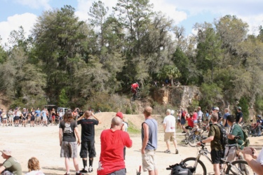 A crowd of spectators watches as a mountain biker performs a jump off a dirt ramp in a wooded area. The audience, consisting of people in casual attire, stands on a dirt surface and holds cameras to capture the action. Lush greenery and trees are visible in the background, and other bicycles are scattered among the crowd. The Vortex mountain bike trail.