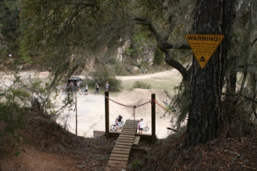 A wooden bridge leads down a slope to a dirt biking area, surrounded by trees and vegetation. In the background, a group of bikers gathers near their bicycles and a vehicle. A yellow warning sign alerts riders to challenging trail features ahead, emphasizing safety precautions. The Vortex mountain bike trail.
