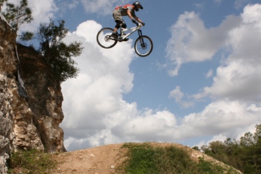 A mountain biker performing a jump off a dirt ramp, with a rocky cliff and a blue sky filled with white clouds in the background. The Vortex mountain bike trail.
