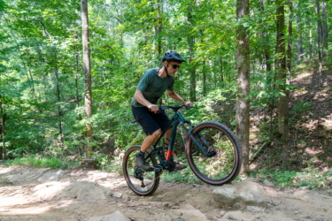 A man in a helmet and sunglasses is riding a mountain bike on a dirt trail, lifting the front wheel over a large rock. He is surrounded by lush green trees in a forested area. Sope Creek mountain bike trail.
