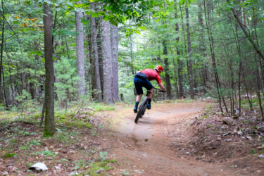 A person riding a mountain bike down a dirt path in a lush forest, surrounded by tall trees and greenery. The cyclist is wearing a red shirt and a helmet, leaning into a turn. Three Sisters mountain bike trail.
