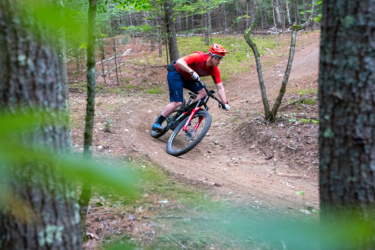 A mountain biker in a red shirt and orange helmet navigates a winding dirt trail in a forested area. The rider leans into a turn, showcasing agility and focus, with trees and underbrush framing the scene. Three Sisters mountain bike trail.