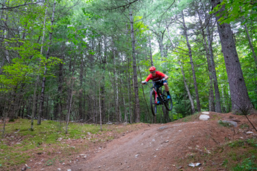 A mountain biker in a red shirt and helmet jumps over a dirt ramp in a forested area, surrounded by green trees and foliage. The rider is airborne, showcasing excitement and skill in a natural trail setting. Three Sisters mountain bike trail.