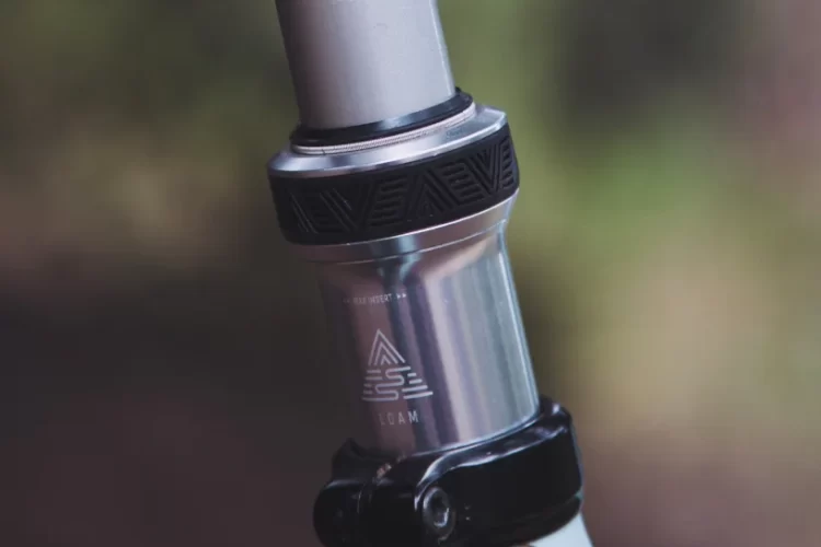Close-up of a mountain bike seat post and clamp, showcasing a silver adjustable mechanism with a black textured ring, attached to a light blue bike frame with subtle patterns. The background is softly blurred, highlighting the bike's components.
