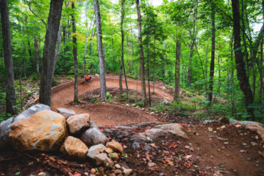 A winding dirt trail curves through a lush green forest, with tall trees and scattered rocks lining the path. A person in a red shirt can be seen riding along the trail, navigating the turn amidst the natural surroundings. Farmer Lake mountain bike trail.