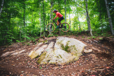 A mountain biker jumps off a large rock in a lush green forest, showcasing agility and skill. The scene captures the thrill of outdoor biking, with trees and fallen leaves surrounding the rocky terrain. Farmer Lake mountain bike trail.