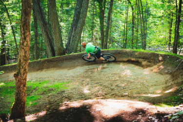 A cyclist in a green shirt and orange helmet leans into a turn while riding a mountain bike along a dirt trail in a lush, green forest. Sunlight filters through the trees, highlighting the curved path and surrounding vegetation. Hiawatha Highlands mountain bike trail.