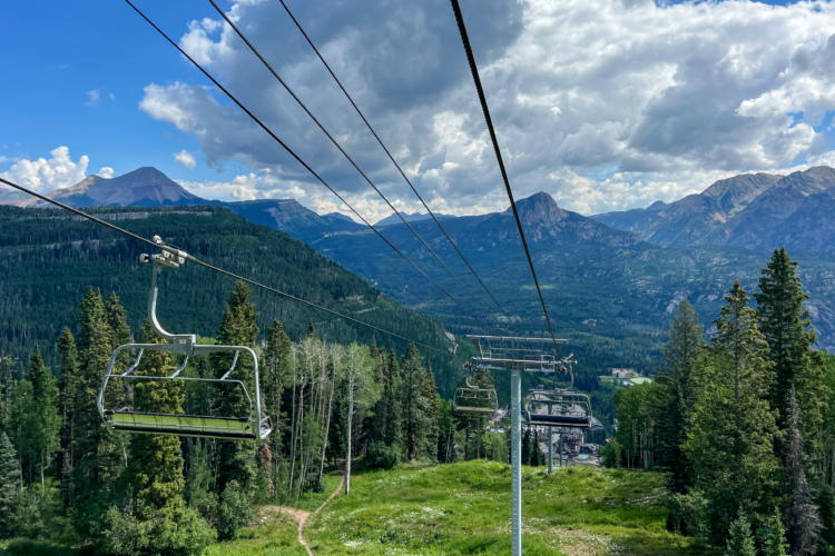 A scenic view from a chairlift overlooking lush green trees and mountains under a partly cloudy sky. The chairlift cables stretch into the distance, leading towards a mountainous landscape with varying elevations.