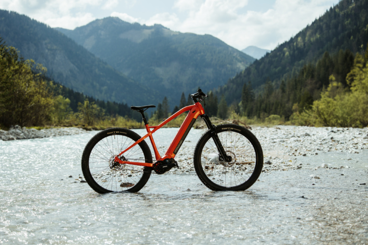 An orange mountain bicycle is standing in shallow water with a rocky riverbed, surrounded by lush green trees and mountains under a partly cloudy sky.