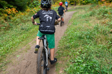 A group of children riding bicycles along a dirt path in a forested area with vibrant autumn foliage. The foreground features a child wearing a black helmet and a black jersey with a graphic design, pedaling a mountain bike. Two other children are visible ahead on the trail, surrounded by greenery and colorful trees in the background. Pinder mountain bike trail.