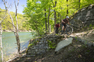 Two mountain bikers navigating a rocky trail beside a lake, surrounded by lush green trees. One rider is ascending a sloped section of the trail while the other appears to be following closely behind. The scene is bright and sunny, showcasing a vibrant, natural landscape. Hobbs State Park Conservation Area mountain bike trail.