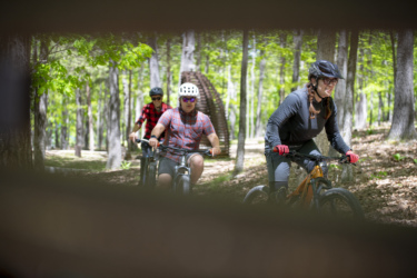 Three mountain bikers riding through a lush, green forest on a sunny day. The first biker, wearing a red flannel shirt, follows slightly behind. The second biker, in a gray plaid shirt and sunglasses, is in the middle, while the third biker, dressed in dark clothing and a helmet with a visor, leads the way. Trees provide a natural backdrop, and a wooden structure is visible in the distance. Hobbs State Park Conservation Area mountain bike trail.