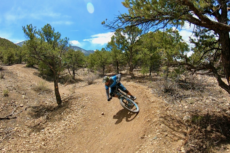 A mountain biker leans into a turn on a dirt trail surrounded by trees and mountains, with a bright blue sky overhead. The biker is wearing a blue jersey and a helmet, showcasing an action-packed moment in outdoor cycling.