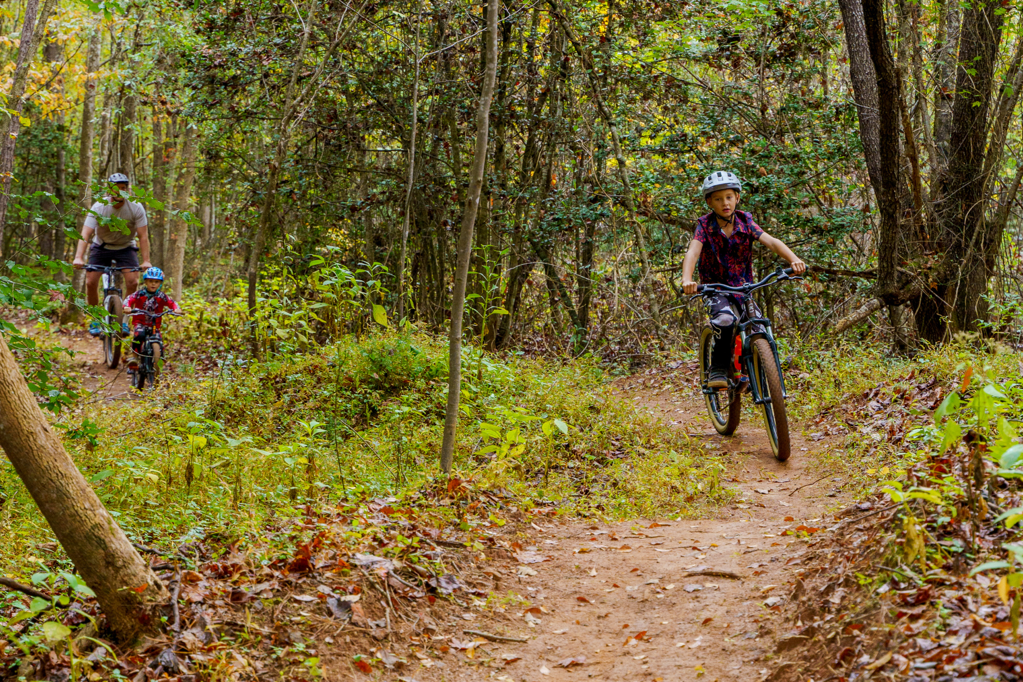 A family of three enjoys a bike ride along a dirt trail in a wooded area. The scene features a child riding a small bike, closely followed by an adult and a younger child. The trail is surrounded by lush greenery and autumn foliage, creating a vibrant and inviting atmosphere for outdoor activities. Everyone is wearing helmets for safety. Oak Hill Community Park and Forest mountain bike trail.