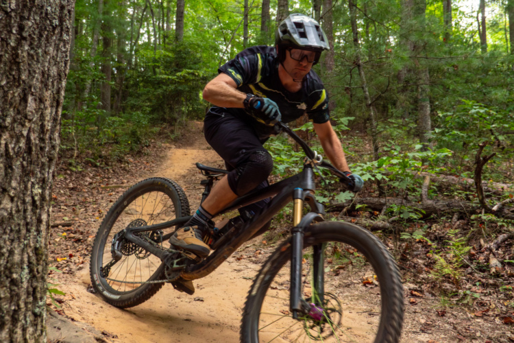 A mountain biker navigating a dirt trail through a lush forest, leaning into a turn while wearing protective gear and a helmet. The scene captures the action and excitement of outdoor cycling amidst greenery and trees.