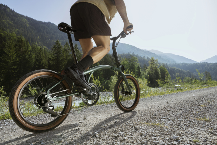 A person riding a green  Brompton G Line folding bike on a gravel path with a backdrop of lush mountains and trees, under a clear blue sky. The cyclist is wearing shorts and cycling shoes, and the scene captures the essence of outdoor adventure and cycling in nature.