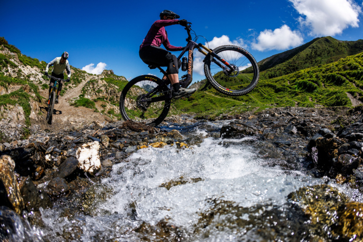 Two mountain bikers navigate a rocky trail in a scenic landscape. One cyclist is performing a wheelie while crossing a stream, with splashes of water visible. The background features lush green hills under a blue sky with some clouds. The environment appears vibrant and outdoor-oriented, highlighting an adventurous moment in mountain biking.