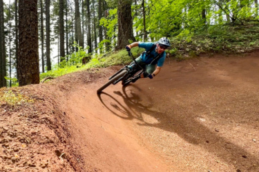 A mountain biker leans into a sharp turn on a dirt trail surrounded by tall trees and lush greenery. The bike's tires stir up dust as the rider maintains balance and control, showcasing a dynamic and energetic moment in outdoor biking. Post Canyon mountain bike trail.