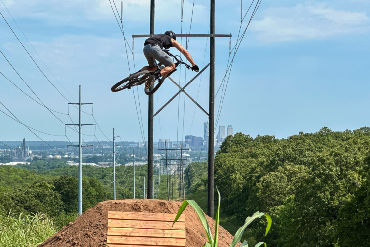 A mountain biker performing a jump over a dirt ramp, surrounded by trees and power lines, with a city skyline visible in the background under a blue sky.