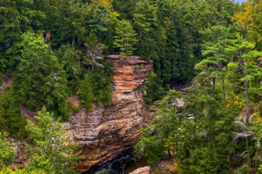 A scenic view of a rocky gorge surrounded by dense greenery, with a river flowing through the canyon. The sky is partially cloudy, and the landscape features various shades of green from the trees, along with rocky cliffs on either side of the water. Ausable Chasm mountain bike trail.